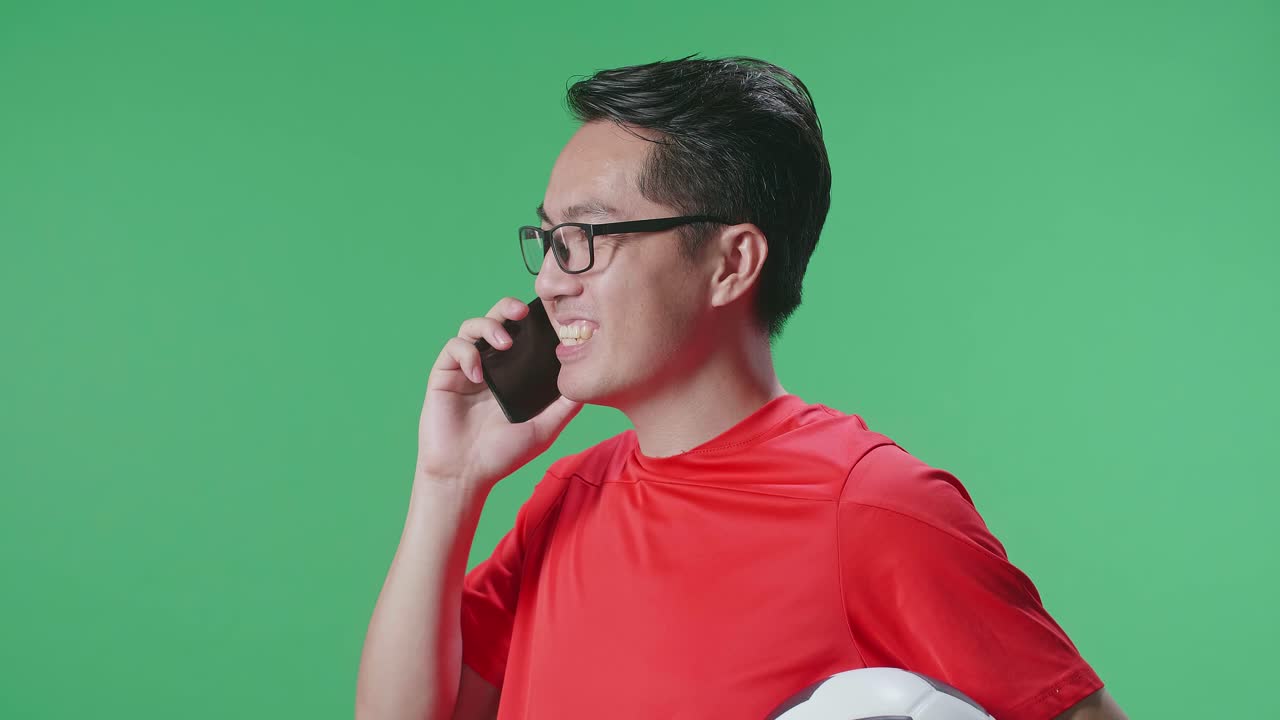 녹색 화면 배경에서 축구를 응원하는 동안 스마트폰으로 대화하는 공을 가진 아시아 남자의 가까운 측면 시각