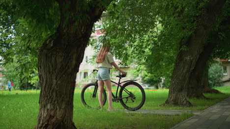 young woman in pink sneakers and jean shorts approaches bicycle on tree-lined path, placing item on seat, background includes lush greenery, building, and person with child running in distance