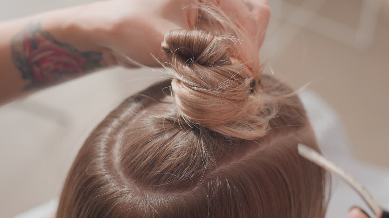 Primer plano de peluquería en un salón: el peluquero carda y sujeta el pelo castaño y rubio dividido en secciones para hacer un moño alto y pulcro, con líneas de partición precisas, utiliza pinzas y horquillas, manos concentradas, estudio suave
