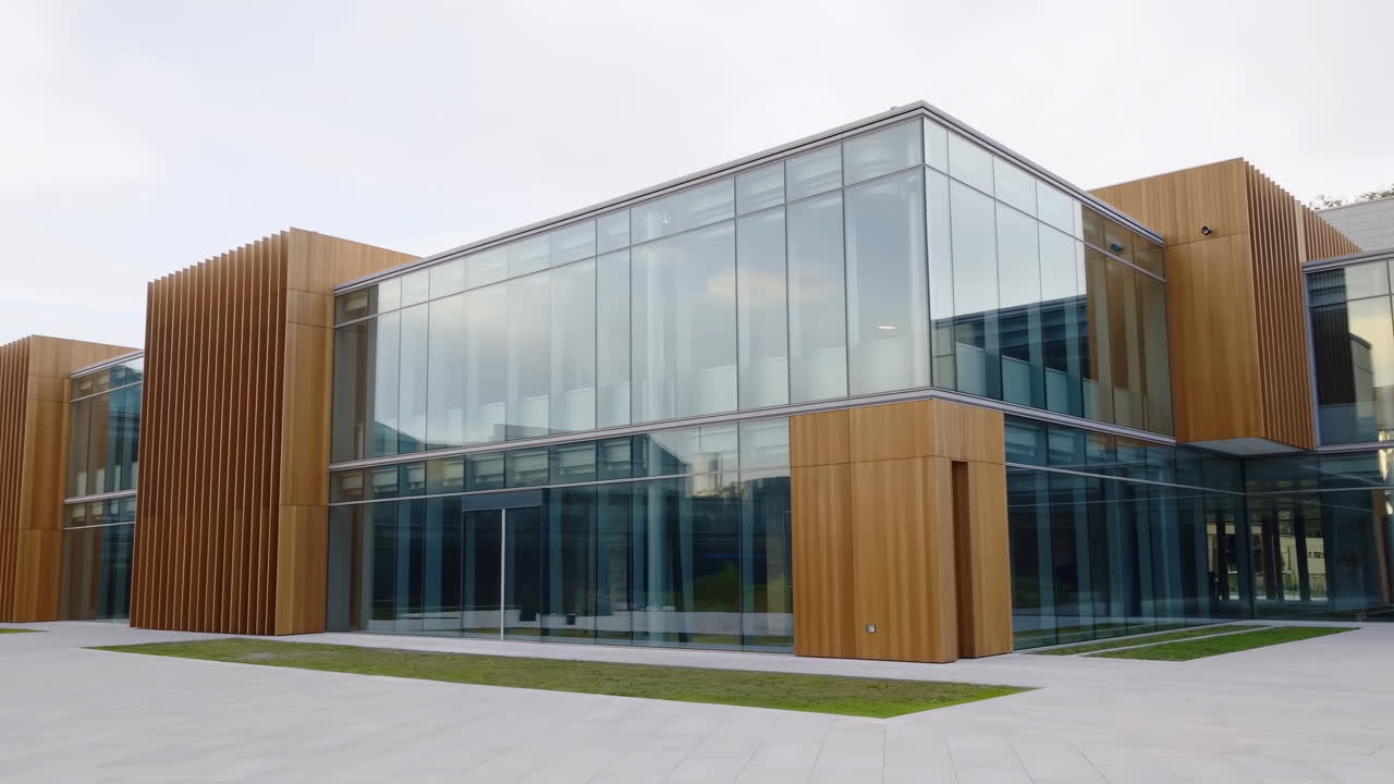 Exterior de moderno edificio de oficinas de cristal, con revestimiento de madera y ventanas de suelo a techo que reflejan el cielo nublado.