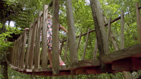 tiro bajo de la espalda de una joven rubia con un vestido largo y colorido de flores cruzando un puente colgante de madera en el bosque