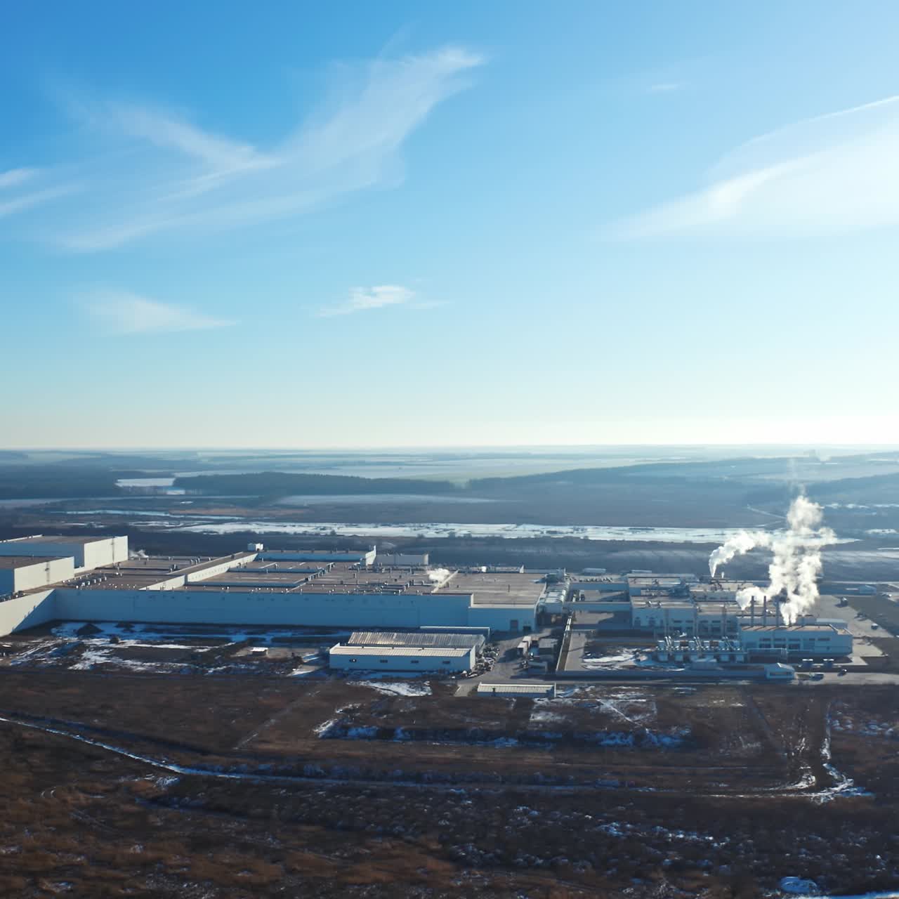 Industrial factory in environment under blue sky. Manufacturing among nature in winter. White smoke coming out of pipes into the air. Aerial view.