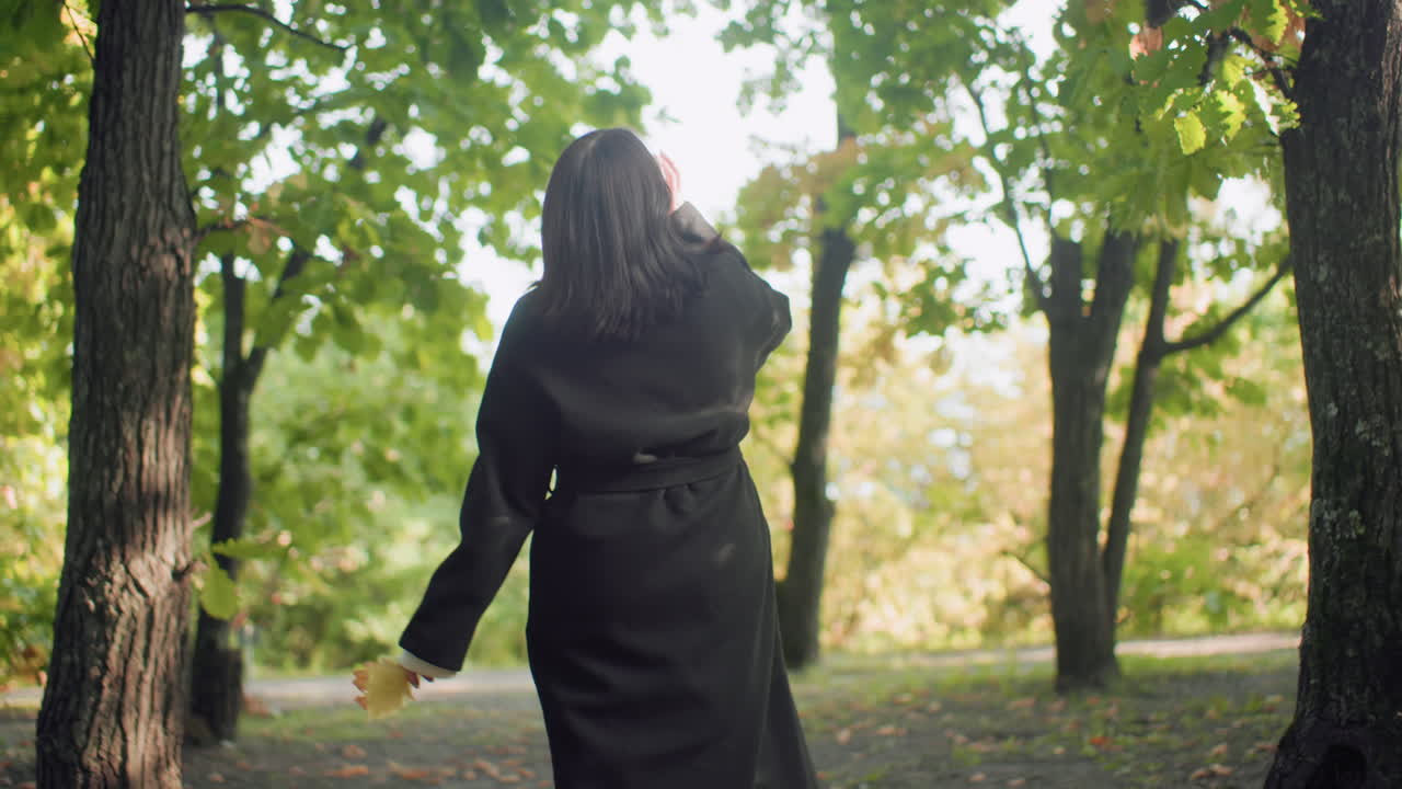 Back view of pensive girl in black jacket strolling under autumn canopy, sipping warm beverage, glancing around between trees, soft sunlight on leaves, relaxed pace and contemplative mood