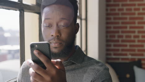 Retrato-De-Un-Joven-Afroamericano-Usando-Un-Teléfono-Inteligente-Tomando-Café-En-Casa-Navegando-Mensajes-En-Red-Enviando-Mensajes-De-Texto-A-Las-Redes-Sociales-Compartiendo-Estilo-De-Vida-En-Línea-Disfrutando-Relajándose-En-Un-Apartamento-Moderno-De-Cerca