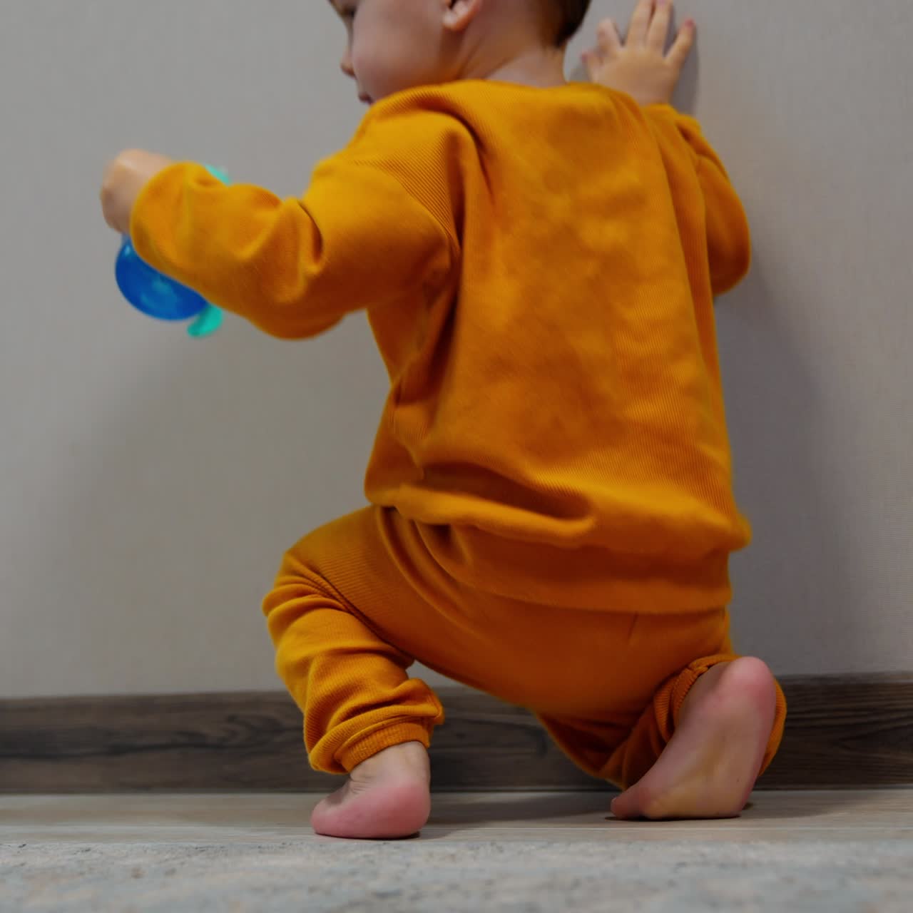One year old boy walks barefoot holding by the wall. Kid stands on toes to reach the bottle and then drops on the floor