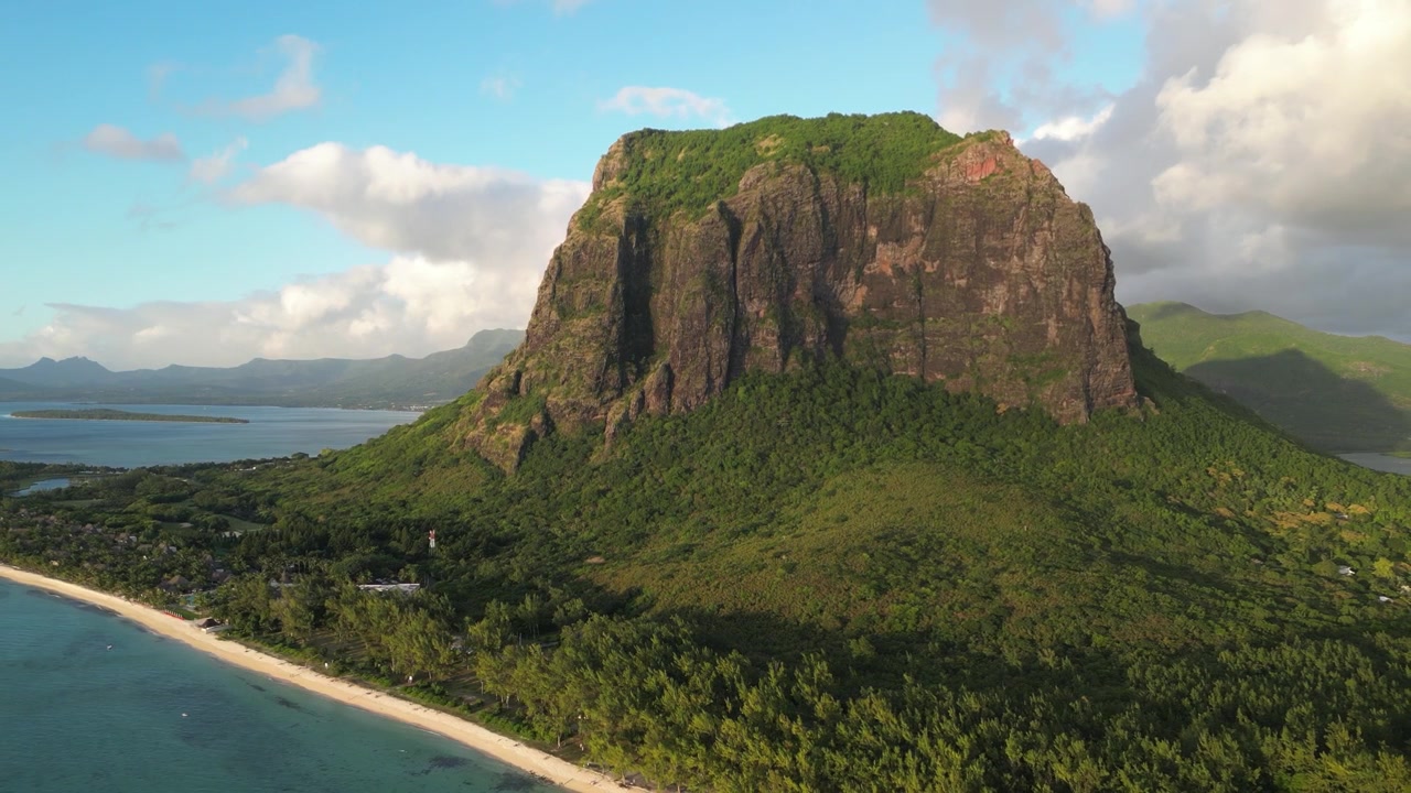 Voo de drone aéreo aproximando-se da enorme montanha Le Morne Brabant nas Maurícias. Monólito basáltico icônico elevando-se sobre a floresta tropical e a costa arenosa. Paisagem de sítio de patrimônio