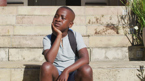 sad african american boy wearing face mask sitting on stairs outdoors on a sunny day