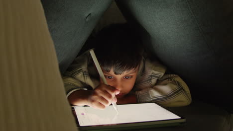 young boy in home made camp made from cushions playing and drawing with digital tablet and stylus at night