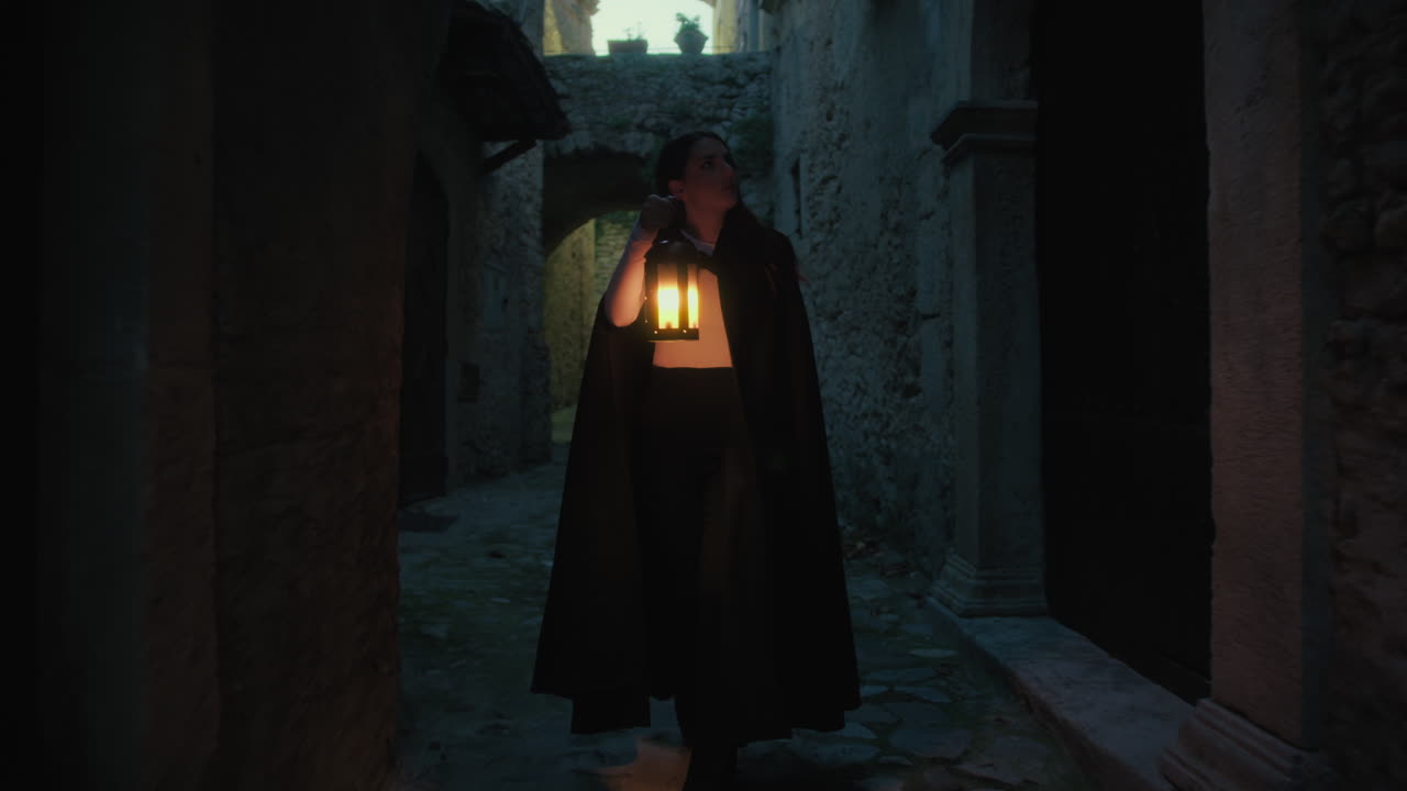 망토와 등불을 입은 여자가 밤에 도시의 거리를 걸어다닙니다.