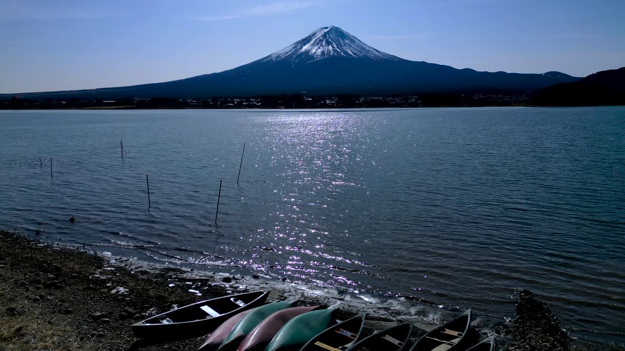 Fuji-bjerget rejser sig over en rolig sø med små både og kajakker, der ligger nær kystlinjen. Fredfyldt japansk landskab, der forener natursceneri med en stille menneskelig tilstedeværelse.