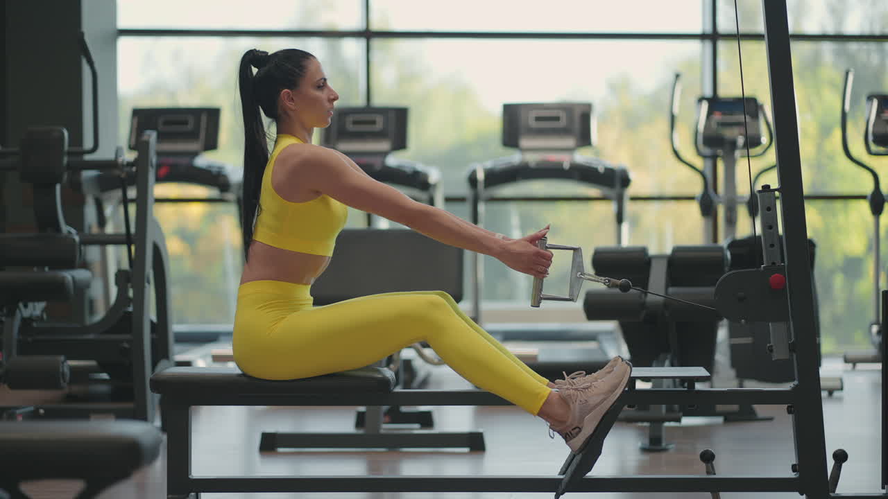 Hispanic woman does exercises on the rowing machine. A girl sits behind a simulator is called a rowing machine. sitting pulls the weight of large Windows and treadmills of the gym