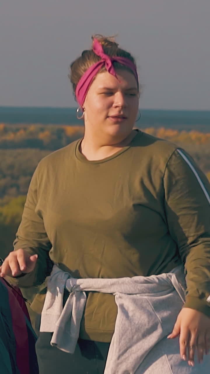 joyful plump tipsy girl dances with raised hands near blue tent against dense autumn forests on river bank slow motion