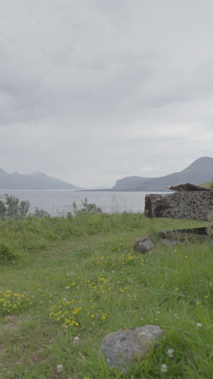 A vertical shot of an abandoned bunker in a grassy field, facing a fjord with mountains under a cloudy sky.