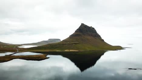 aerial view of the majestic mount kirkjufell reflecting in the calm lake, surrounded by green fields and a winding road, showcasing the breathtaking beauty of iceland's natural landscape