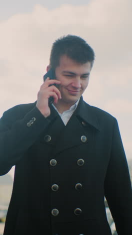 freelancer con abrigo negro sonriendo y hablando por teléfono con emoción mientras hace gestos en una vibrante área urbana con edificios modernos de vidrio, coches estacionados, luz solar suave y atmósfera alegre