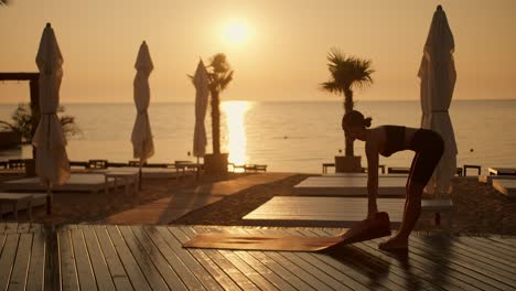 A-brunette-girl-in-a-black-sports-summer-uniform-spreads-a-mat-before-meditation-and-yoga-classes-on-a-Sunny-beach-in-the-morning.-Sunny-Bronze-colored-beach-with-palm-trees-covered-with-boards