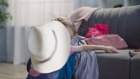 little girl plays with hat putting into suitcase on sofa