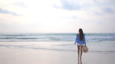 back to the camera, slow motion, rule of thirds, an attractive woman in shorts and a bohemian blouse holds a sunhat in one hand as she walks into the oncoming surf