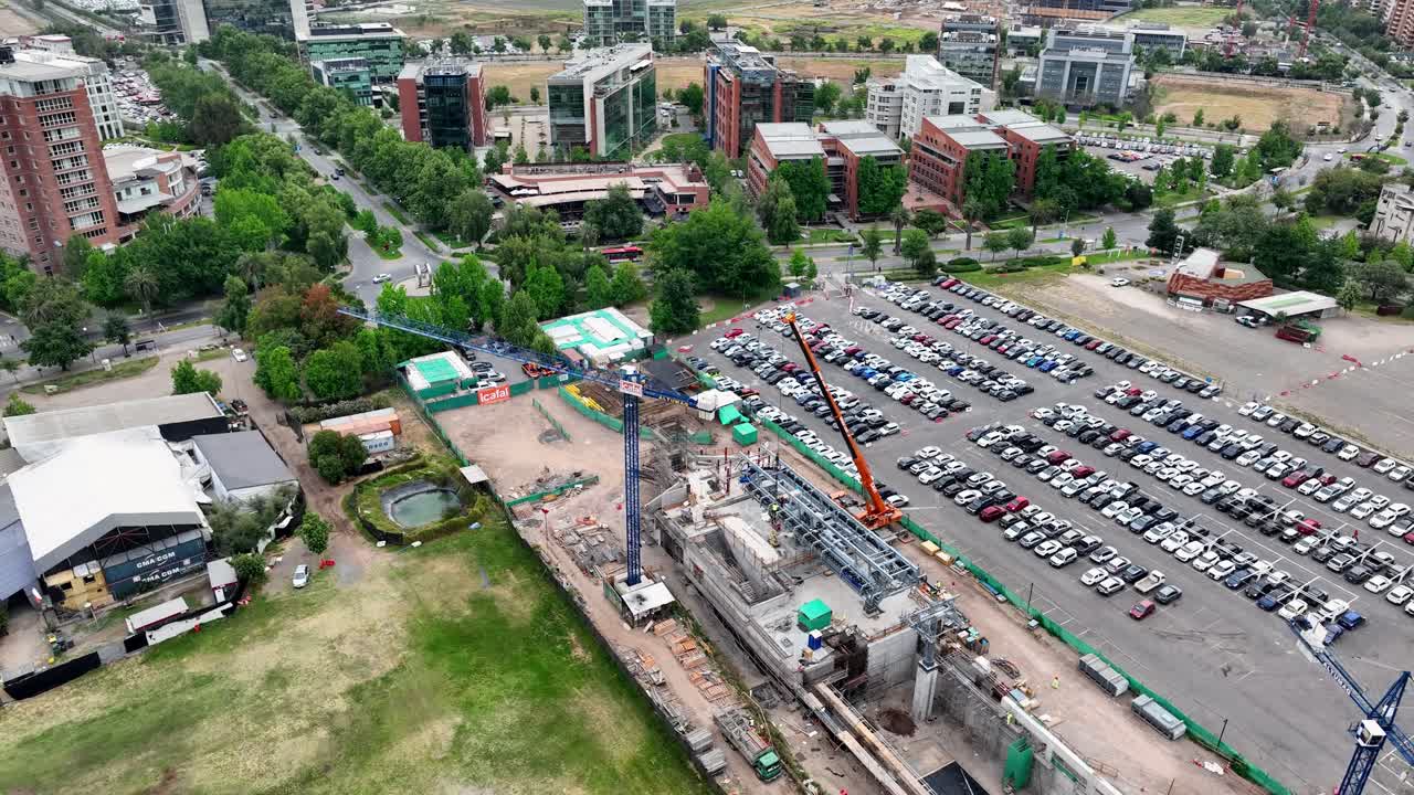 Orbiting drone aerial of urban construction site with colorful cranes, large parking lot with organized car rows, and city buildings