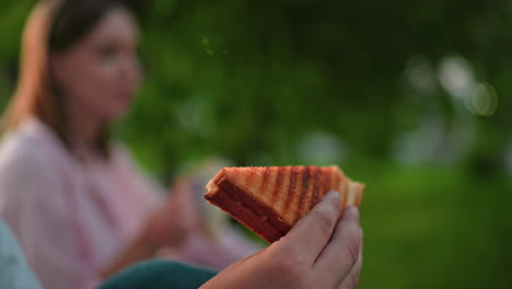 vista parcial de cerca de una persona sosteniendo un sándwich a la parrilla con un fondo ligeramente borroso de personas comiendo, al aire libre, entorno natural con iluminación suave