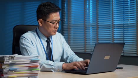 Focused Professional Engaged in Work at Night with Laptop in Office Surrounded by Stacks of Paperwork and Soft Blue Lighting Creating a Productive Atmosphere
