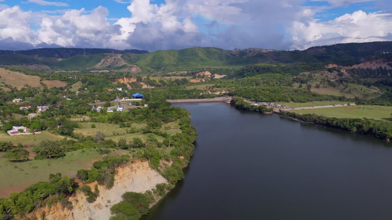 Aerial view of López–Angostura reservoir and dam near Santiago, Dominican Republic, surrounded by green hills, farmland and calm water under dramatic tropical clouds. Wide shot