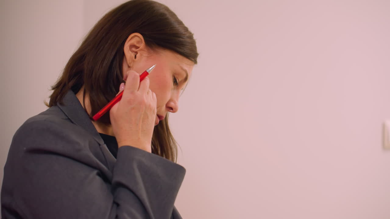 Professional woman in business attire holding pen, thoughtfully gazing off to the side. She seems deep in thought while adjusting pen, side profile view, neutral background setting