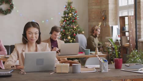 Oung-Pretty-Office-Manager-In-Smart-Casualwear-And-Headset-Networking,-Her-Colleagues-Are-Working-Sitting-On-Desk-In-Background