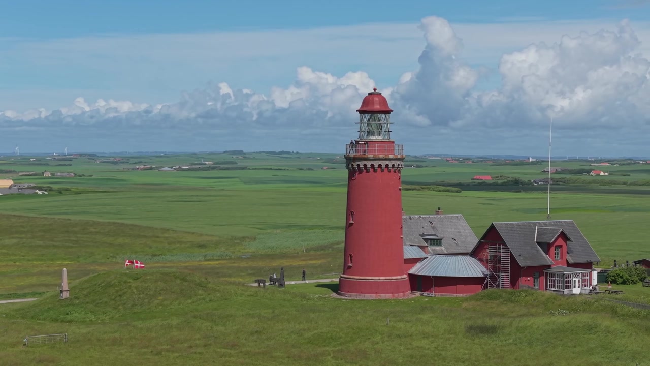 Luftaufnahmen, die den Leuchtturm Bovbjerg im Herzen einer farbenfrohen westjütländischen Landschaft umkreisen, mit grünen Feldern, blauem Himmel und ziehenden weißen Wolken