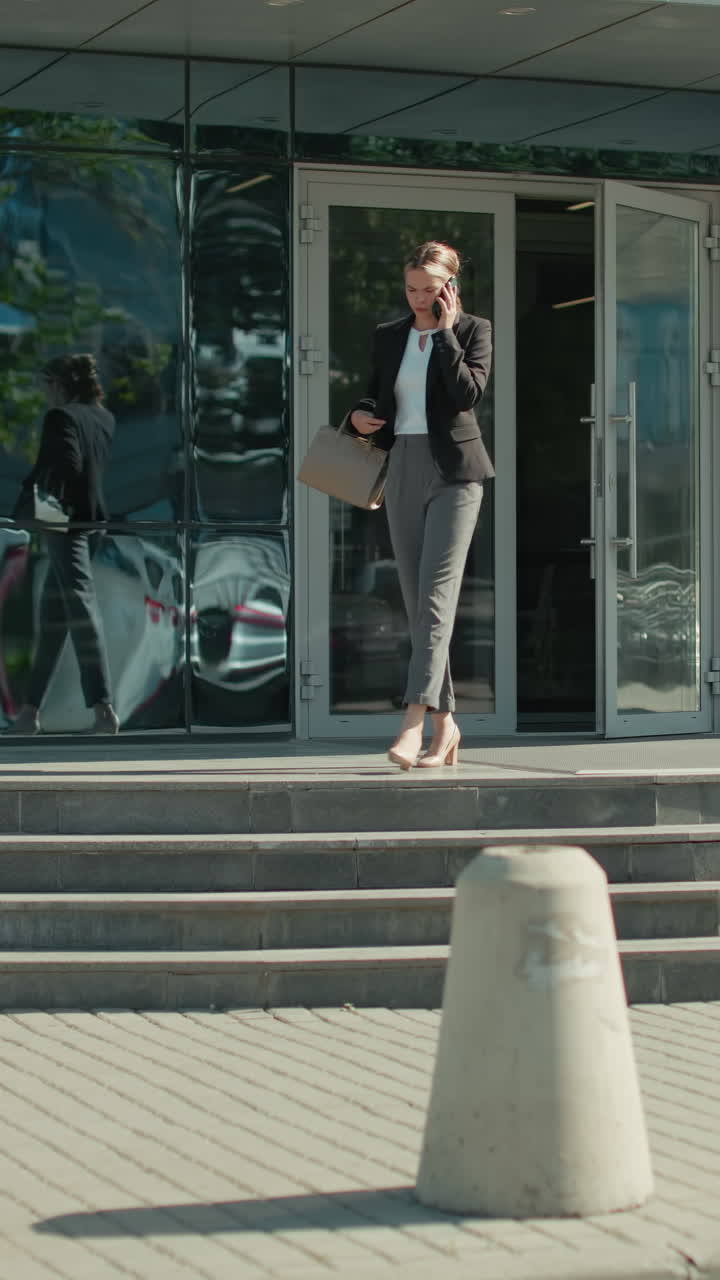 Confident businesswoman exiting modern office building talking on phone after client meeting, wearing formal attire with shopping bag in hand