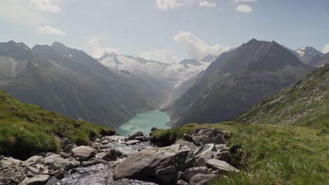 hay un pequeño arroyo que fluye en primer plano y los alpes austriacos, así como un hermoso lago azul al fondo.