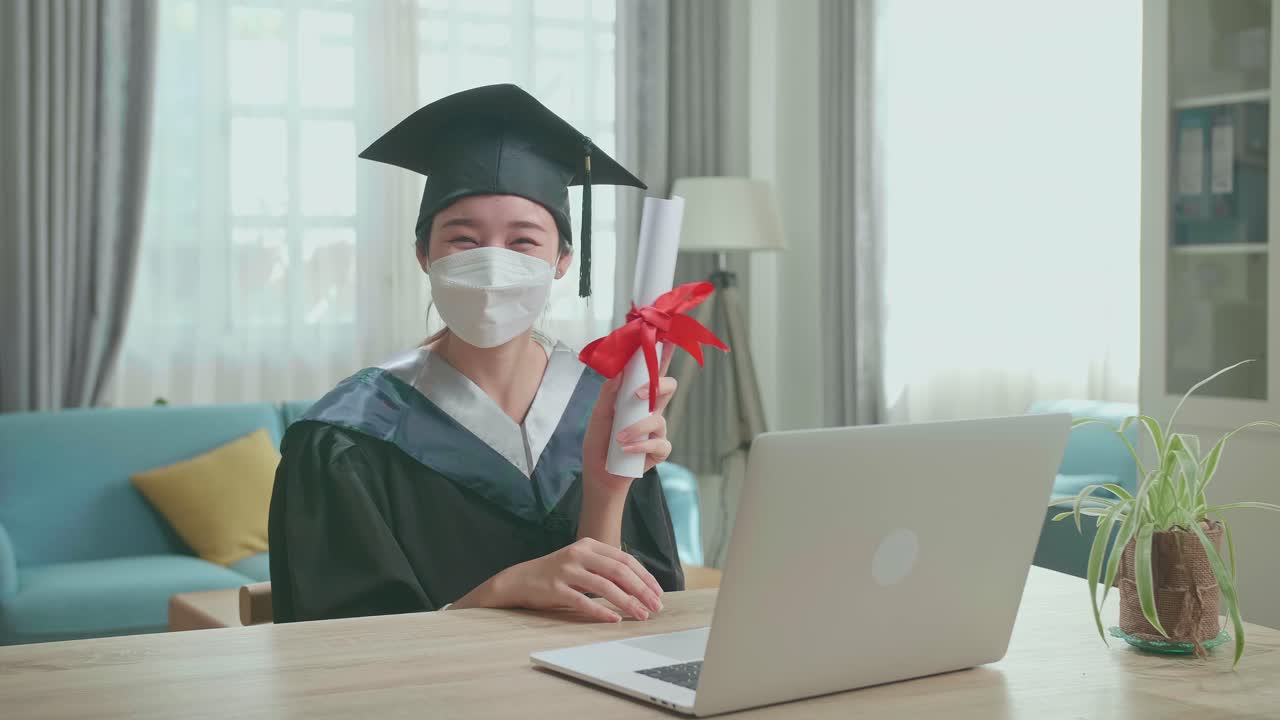 Excited Asian Woman Wearing Protection Face Mask And Holding A University Certificate, Smiling To Camera. Pretty Female Graduate Wearing A Graduation Gown And Cap Sitting On The Living Room