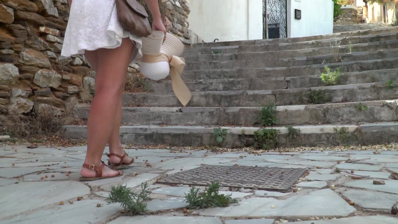 siguiente plano de piernas femeninas con vestido de verano y sandalias sosteniendo un sombrero subiendo escalones en un callejón griego