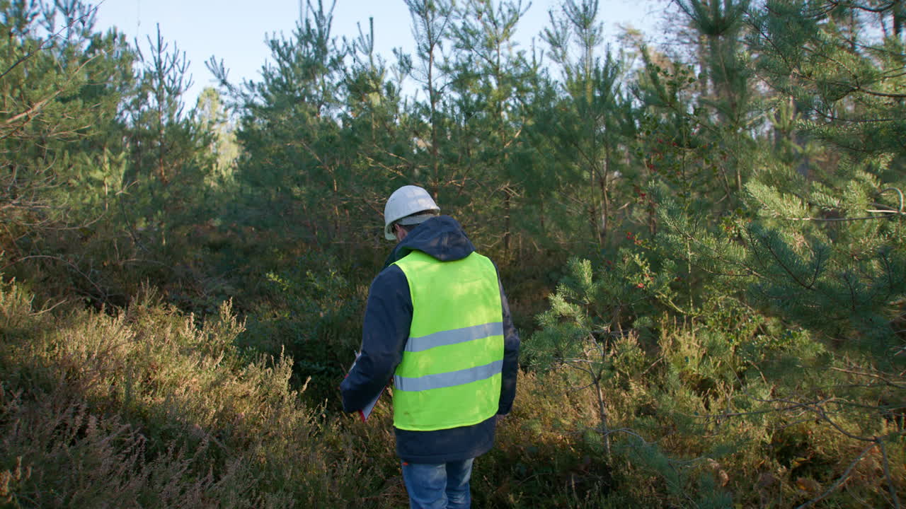Male biologist walking over tall grass while looking around at the forest while carrying a clipboard, handheld