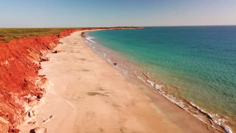 Aerial-footage-of-male-and-female-couple-sitting-atop-red-cliff-overlooking-beach-and-blue-ocean