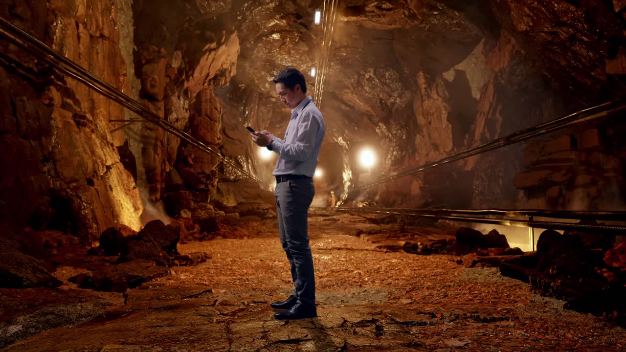 Full Body Side View Of An Asian Male Professional Worker Standing With His phone In Underground Mine Tunnel, Working Continuously With His phone