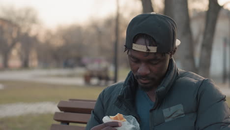 Close up of young man in cap and puffer jacket on park bench looking down, eating fish pie during calm evening, bokeh background, winter trees, relaxed solitary snack in quiet urban setting