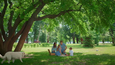 young family resting with dog in shadow on weekend. parents lunching with kids.