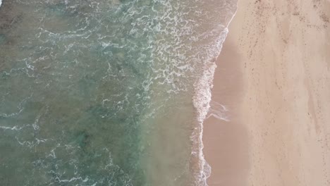 Vista-Aérea-Superior-Sobre-El-Agua-Clara-Y-Turquesa-Del-Océano-Que-Hace-Espuma-En-La-Orilla,-Lavando-La-Arena-Dorada-De-La-Playa-Con-Huellas,-Vacaciones-De-Verano-En-La-Isla-De-Hawaii