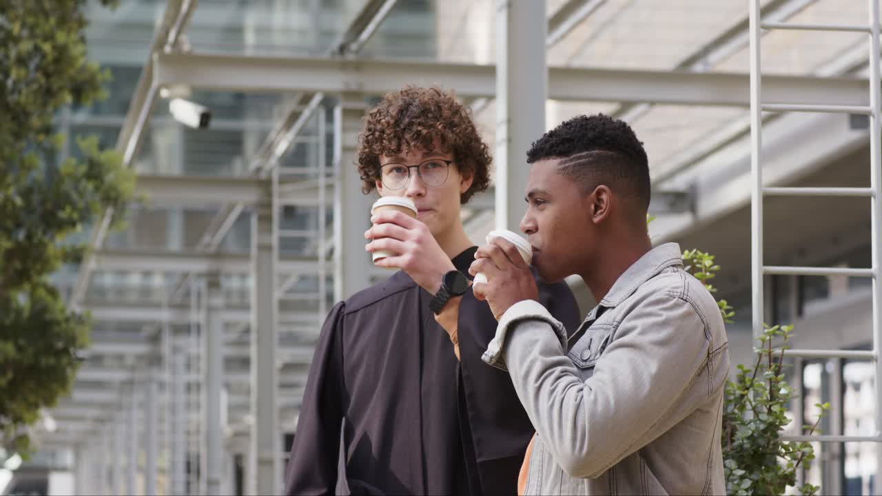 Laureato maschio bianco che accetta un caffè da un amico con la giacca di jeans sotto un pergolato fuori dal campus, conversando.