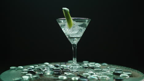 Rotating-martini-glass-with-a-slice-of-lemon-and-ice-blocks,-rotating-on-a-beautiful-plateau-with-shiny-stones,-shot-on-a-black-infiniy-background