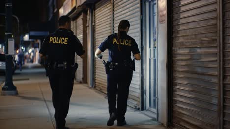 Two police officers walk along a quiet city street at night, checking for any suspicious activity. They remain alert while providing safety to the local residents.