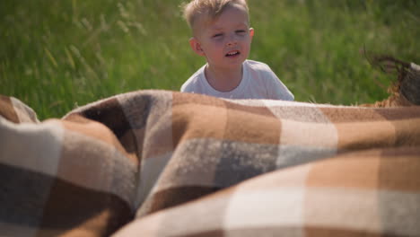 a young boy in a white shirt smiles as he holds onto the edge of a flowing plaid scarf in a grassy field, with the wind gently blowing. the boy looks up at someone in the background