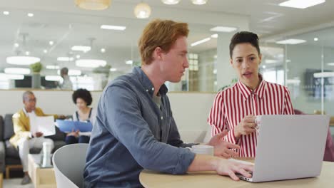 Diverse-male-and-female-colleagues-sitting-at-desk,-using-laptop,-having-business-talk-in-office