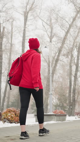 view of athlete walking along patterned pathway with bag over shoulder touching chest, surrounded by snow-dusted benches, trees, light poles, and distant building in serene foggy atmosphere