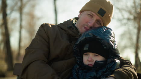 father and son sitting on a bench, with the father head resting on his son's head, with the father holding his son close, both look focus, with blurred view of trees in the background