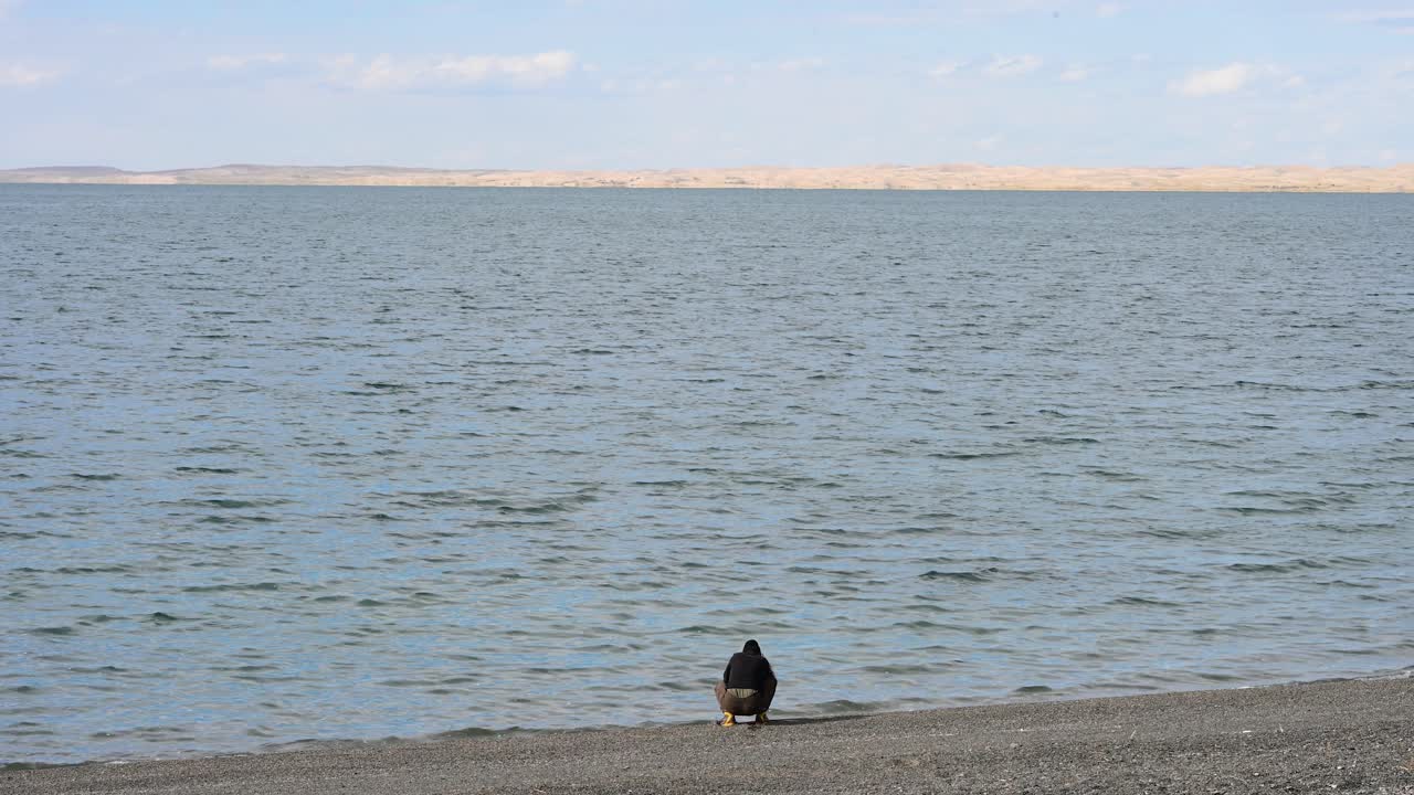 A woman kneels at the water's edge of Durgun Nuur lake in Mongolia, using her smartphone to capture the scenic view. A moment of modern tourism in a remote landscape