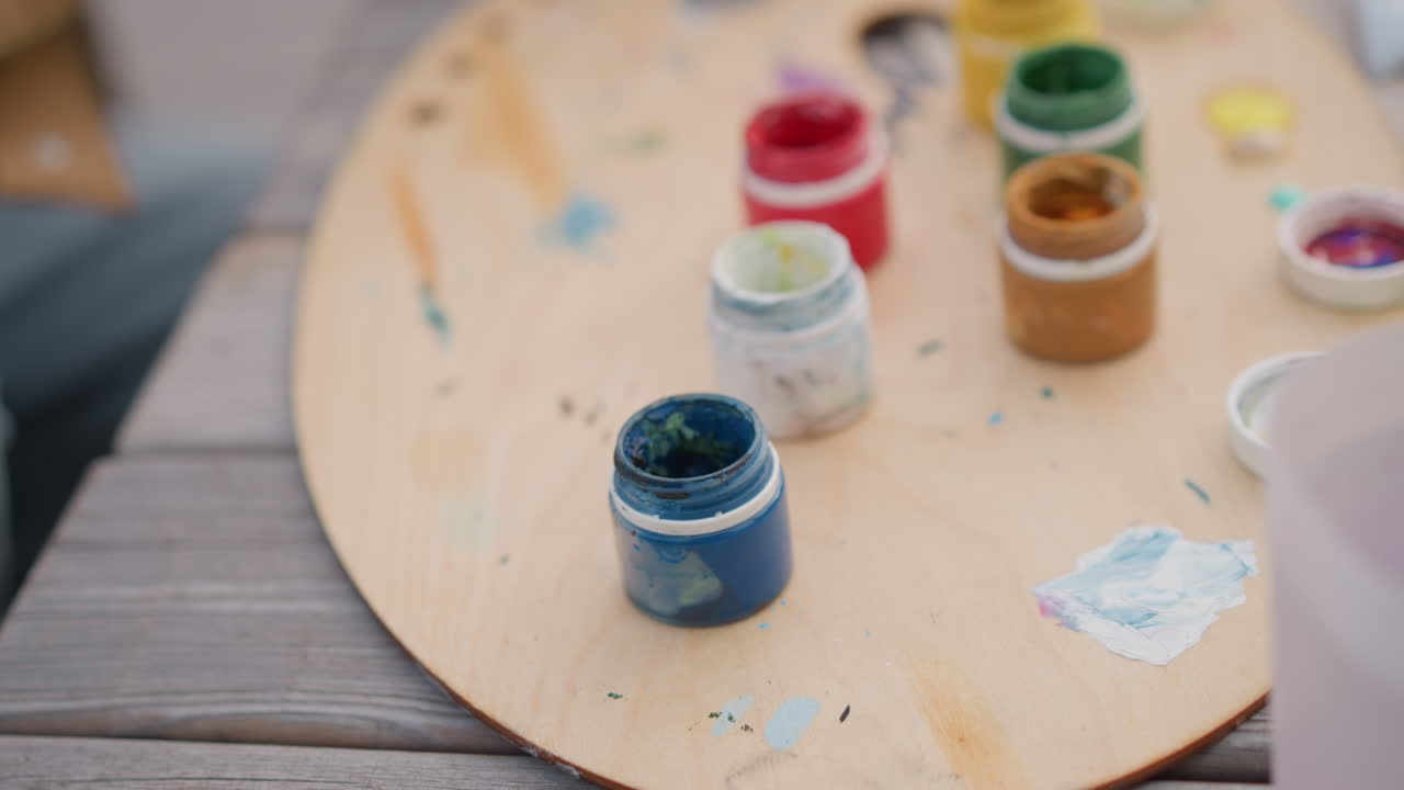 mani che aprono un barattolo di vernice blu, palette di legno e pennelli nelle vicinanze, pittore che sistema i pigmenti acrilici su un tavolo rustico, focus ravvicinato sulla rimozione del coperchio e sul pigmento materico, atmosfera creativa e tranquilla con strada