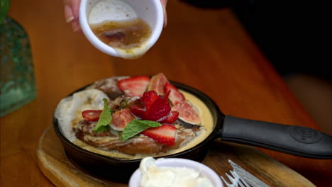 slow motion of a person pouring a fruit sauce onto a cinnamon roll topped with sliced figs and strawberries in an iron skillet