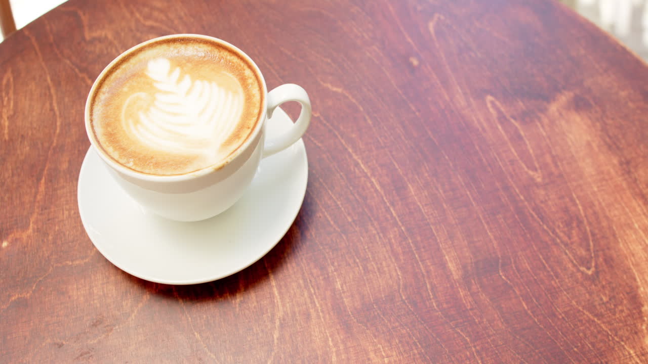 A cup of cappuccino with latte art sits on a wooden table in a cafe, with copy space